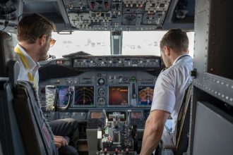 Dois pilotos operam os sistemas e instrumentos de um cockpit de avião comercial.