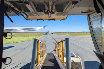 Vista do pátio de um aeroporto a partir da porta de carga de uma aeronave, com outros aviões ao fundo.