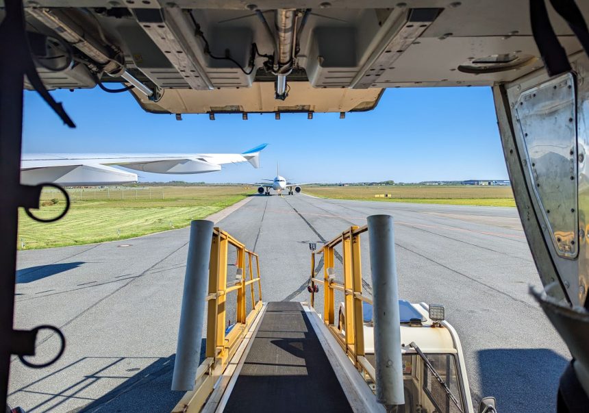Vista do pátio de um aeroporto a partir da porta de carga de uma aeronave, com outros aviões ao fundo.